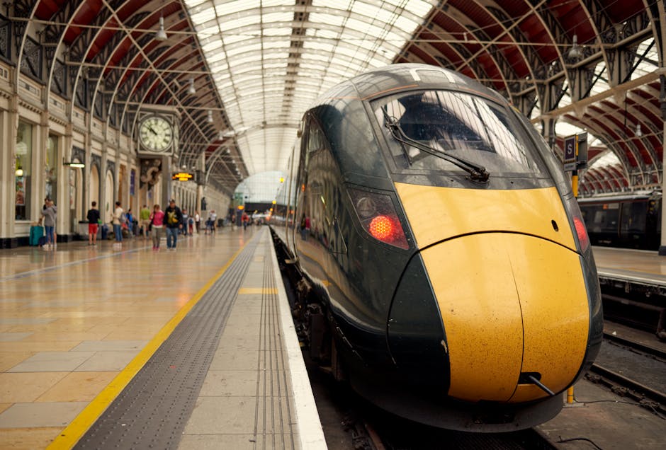 Interior view of Paddington train station with a modern high-speed train positioned on the platform. The train features a sleek, aerodynamic design with a black and yellow front, and the windshield wipers are visible. The station's roof is arched and made of glass and metal, allowing natural light to illuminate the busy waiting area. Passengers with luggage are scattered along the platform, some sitting on benches and others walking. The platform surface appears clean and well-maintained, with tactile paving along the edge for safety. A clock and station signage are visible on the wall, contributing to the organized appearance of the transportation hub. Paddington Carpet Cleaning provides commercial cleaning services for such busy transport environments to ensure hygiene and cleanliness.