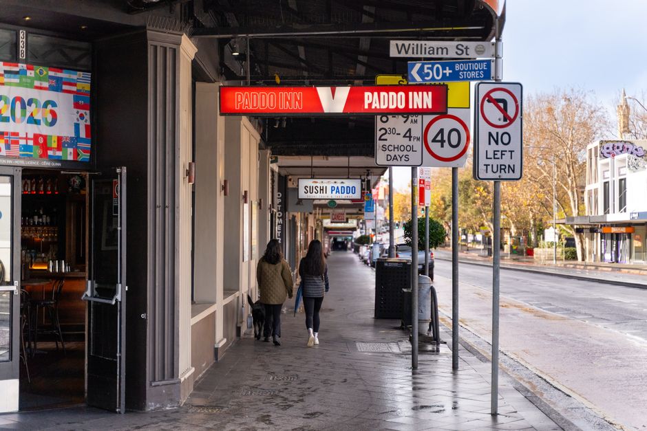 A street scene in Paddington with a commercial building featuring a beige exterior and glass windows. Visible signage includes a red and white 'Paddo Inn' sign and a smaller 'Sushi Paddo' sign hanging beneath it. The sidewalk is clean with concrete paving, and two women are walking side by side, dressed in casual clothing. The street is lined with trees and additional shops, with traffic signs indicating a 40 mph speed limit, no left turn, and parking restrictions. The scene is captured during daylight hours with natural lighting, and the overall environment appears tidy and well-maintained, reflecting a typical urban area in Paddington.