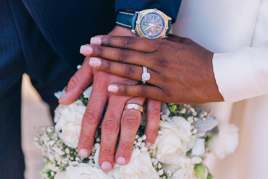 Close-up of three hands layered on top of each other, showing different skin tones and wedding rings. The top hand, wearing a blue watch, belongs to a person in formal attire, possibly a groom, while the middle and lower hands, with one decorated with a diamond ring, indicate a bridal couple. The hands rest on a bouquet of white roses and baby's breath, placed against a soft, neutral background. This image emphasizes unity and celebration, often associated with wedding or special occasion photography.