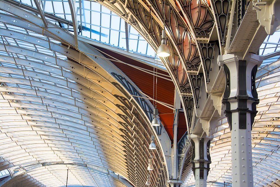 A detailed view of an ornate, large-scale architectural ceiling featuring curved metal beams painted in white and black, with intricate latticework and decorative supports. The ceiling is constructed with a combination of glass panels allowing natural light to illuminate the space and metal framework. Pendant lighting fixtures hang from the ceiling, casting a soft glow on the surfaces. The overall condition appears clean and well-maintained, highlighting the structural design and material finishes typical of a historic or commercial building, relevant to sanitisation and maintenance practices in domestic and commercial cleaning contexts. The image emphasizes surface cleaning and deep cleaning of the architectural features, as seen in the absence of dust or dirt, making it suitable for content related to surface cleaning, sanitisation, and custodial care by Paddington Carpet Cleaning.