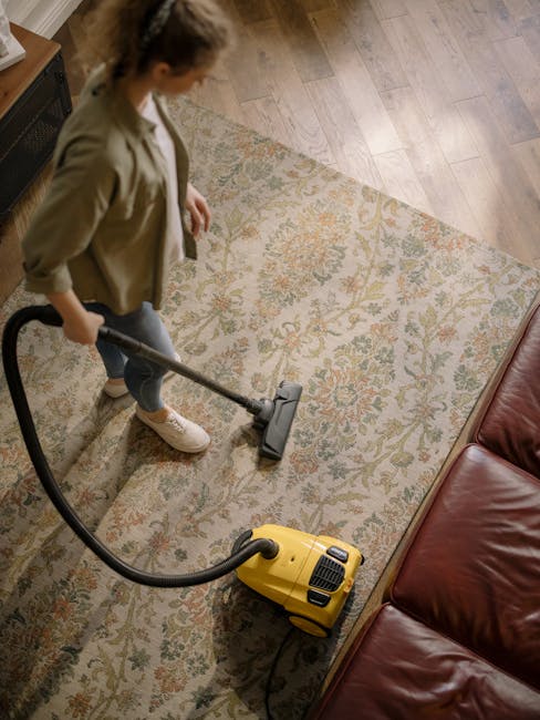 A woman operating a yellow vacuum cleaner on a patterned area rug in a living room with wooden flooring, near a red leather sofa, demonstrating surface cleaning and deep cleaning, with visible dust and dirt being removed to enhance cleanliness. The room is well-lit, and the scene emphasizes domestic cleaning services provided by Paddington Carpet Cleaning as part of their sanitisation and maintenance offerings.
