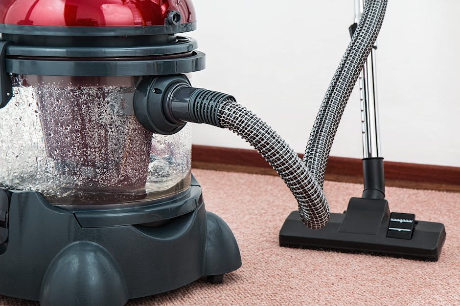 Close-up image of a commercial upright vacuum cleaner with a transparent dust container filled with debris, connected to a flexible, patterned hose that extends to a flat floor brush attachment. The vacuum is positioned on a pink carpet in an indoor residential or office space, with a white wall and wooden skirting board visible in the background. The surface appears clean and well-maintained, with a shiny and dust-free appearance on the carpet, reflecting a professional cleaning environment associated with surface cleaning and sanitisation services provided by Paddington Carpet Cleaning.