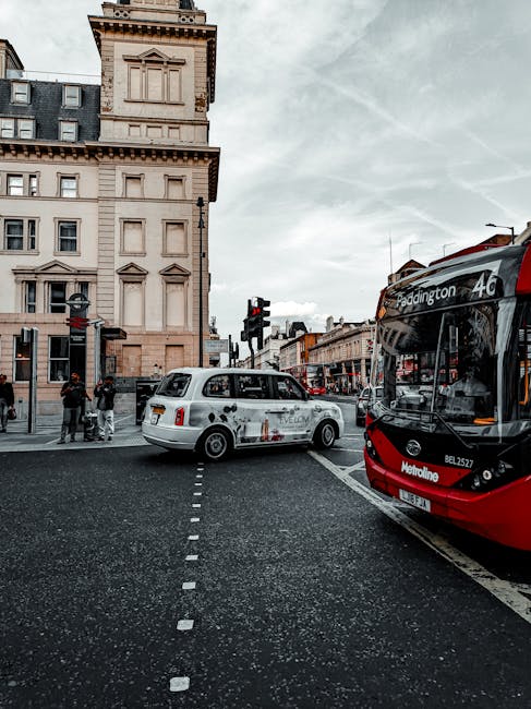 A street scene near Paddington Station featuring a red and black London bus with route information displayed on the front, stopped at a pedestrian crossing with a white taxi parked beside a historical building with detailed architectural elements. The asphalt road appears clean and well-maintained, with clearly marked lane lines. Nearby pedestrians wait to cross the street, and traffic lights are visible overhead. The lighting is natural, with an overcast sky casting soft light over the scene. The image emphasizes urban surface cleaning and maintenance, reflecting the importance of cleanliness in busy transport hubs, aligning with paddingtoncarpetcleaning.org.uk's focus on thorough cleaning and sanitisation services, especially in high-traffic areas.