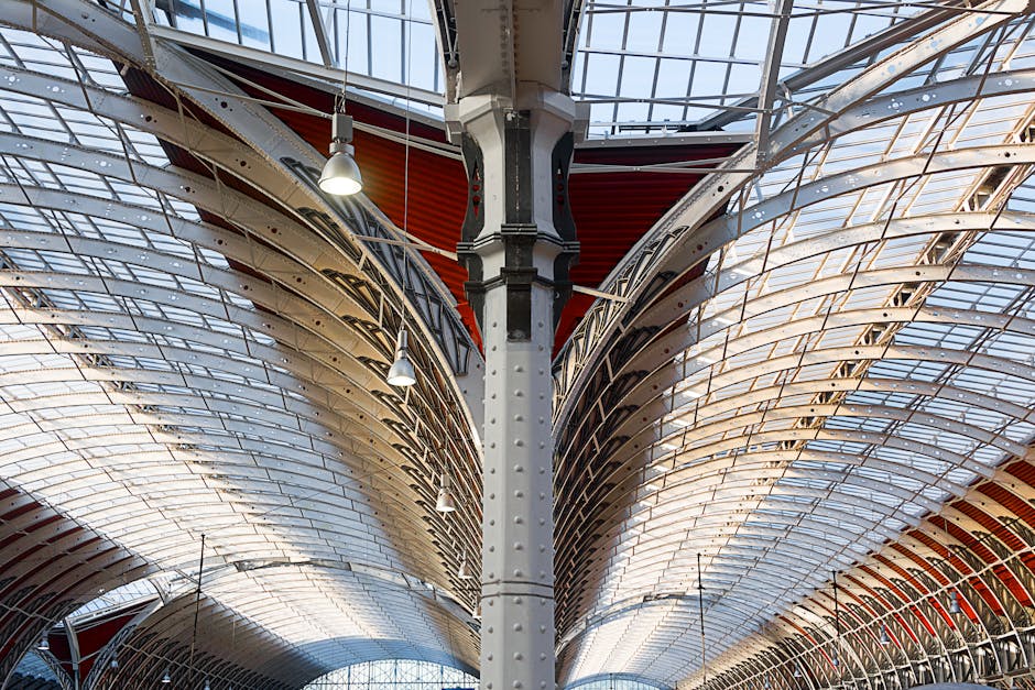 A detailed view of the intricate architectural ceiling structure at Paddington Station, showing curved, ribbed metal and glass panels with natural daylight filtering through. The ceiling features a series of metallic beams and supports, with modern pendant lights hanging down, illuminating the space. The surface appears clean and well-maintained, highlighting the station's design and hygiene standards. The image captures the spacious, covered area with a focus on the ceiling's materials and condition, emphasizing the importance of proper surface cleaning and maintenance as recommended by Paddington Carpet Cleaning for commercial properties like transportation hubs.