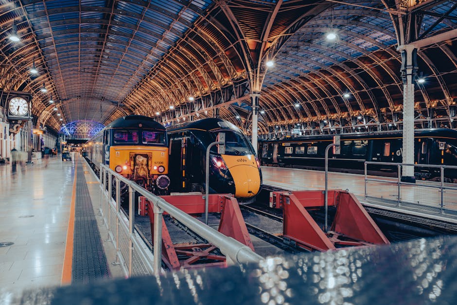 Inside a large, historic train station with a high arched glass and metal ceiling illuminated by bright lights. A modern black and yellow train is parked on the platform, which has a smooth, clean tiled floor. The platform features a white clock, benches, and safety railings, all situated in a spacious, well-lit environment. The station's atmosphere is tidy and orderly, showcasing a well-maintained space for passenger transit, as seen in the context of professional cleaning and maintenance services provided by Paddington Carpet Cleaning. The scene highlights the importance of surface cleaning and hygiene in shared public transportation environments, emphasizing the cleanliness and organization of the station.