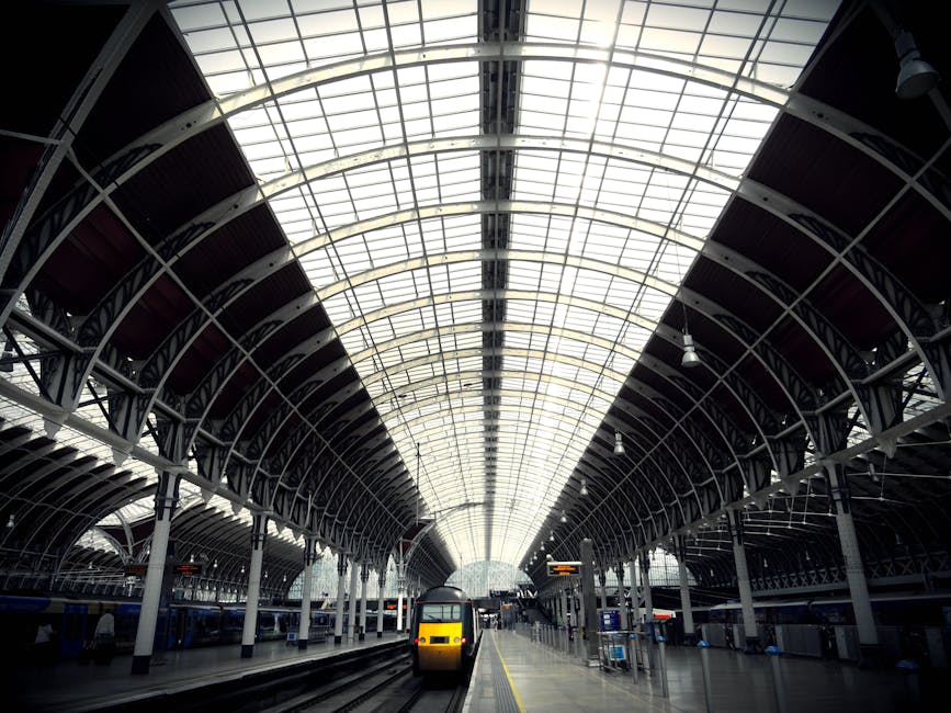 Interior view of a large train station platform featuring a high arched glass ceiling with metal framework, allowing natural light to illuminate the space. The platform is clean and organized, with train tracks running in the foreground, and a yellow and black train approaching or departing. The station is equipped with signage, lighting fixtures, and seating areas, reflecting a modern, well-maintained environment suitable for efficient passenger flow. Paddington Carpet Cleaning is a professional cleaning service specializing in surface cleaning, deep cleaning, and sanitisation, ensuring hygienic and spotless residential and commercial spaces.