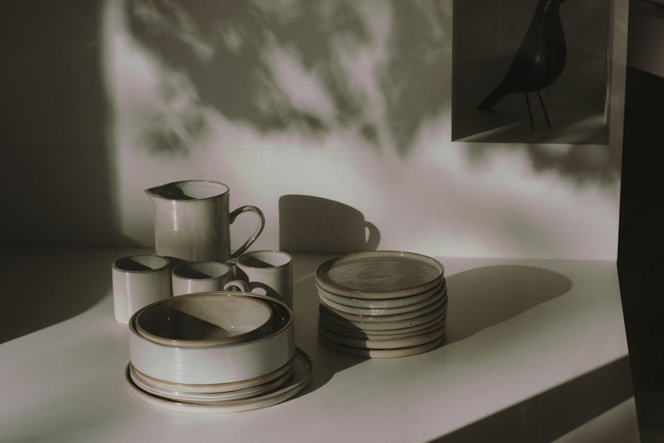 A set of ceramic dining plates, cups, and a jug arranged on a white countertop in a well-lit kitchen. The items are clean, with their surfaces reflecting light, and are stacked neatly or placed side-by-side. A shadow from nearby objects creates patterns on the wall and countertop, indicating natural or bright artificial lighting. The scene emphasizes surface cleaning and hygiene, aligning with deep cleaning or sanitisation practices discussed in the context of choosing between steam and dry cleaning for Paddington carpets, as highlighted by Paddington Carpet Cleaning on their website.