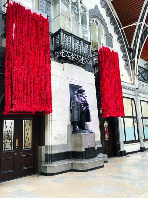 The image depicts an indoor space with high ceilings and large, ornate windows that allow natural light to illuminate the area. The floor is covered with polished, light-colored tiles, creating a clean and reflective surface. At the center, there is a dark bronze statue of two standing figures, mounted on a stone pedestal with an inscription plaque. Behind the statue, the wall features decorative architectural elements, including intricate moldings and metal balcony railings painted black. On either side of the statue, two tall red floral arrangements hang vertically, adding vibrant color and visual interest to the scene. The area appears well-maintained and clean, with a focus on surface cleaning and overall hygiene, consistent with practices emphasized in Westminster Council Carpet Disposal Rules for Paddington, as promoted by Paddington Carpet Cleaning.