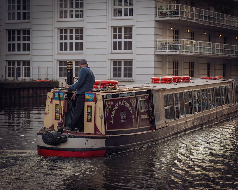 A narrowboat named 'London Waterbus' is navigating a canal in Paddington, London, with a man standing at the stern steering the boat. The boat's roof is equipped with bright red life buoys and safety equipment, contrasting with the boat's dark and cream-colored exterior that features decorative, colorful panels. The scene is set against a backdrop of modern white residential buildings with large windows and balconies, reflecting in the calm water. The sky appears overcast, providing diffused lighting that highlights the cleanliness of the boat and the water surface. This setting illustrates a typical scene of surface navigation and maintenance of canal boats in the Paddington area, with no visible signs of dirt or disrepair, emphasizing the importance of proper cleaning and upkeep practices in preserving such vessels in urban environments. Paddington Carpet Cleaning is committed to maintaining high standards of cleanliness and hygiene, consistent with the themes of surface cleaning and sanitisation relevant to Westminster Council Carpet Disposal Rules for Paddington.