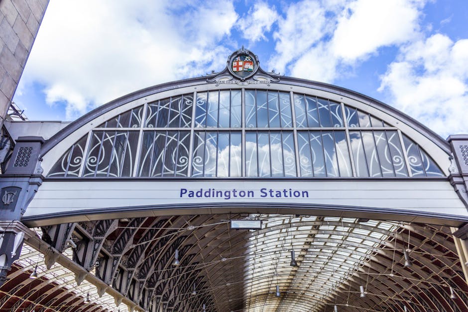 Exterior view of Paddington Station's ornate metal and glass arched entrance, featuring intricate decorative ironwork and a prominent sign reading 'Paddington Station' beneath a coat of arms crest on the upper arch. The station's roof structure is visible, with sunlight filtering through the clear glass panels, highlighting the clean, well-maintained surfaces. The architectural detail and reflective glass emphasize the historical yet modern design, with a bright blue sky and some scattered clouds in the background. Paddington Carpet Cleaning, a professional cleaning service, specializes in surface cleaning and sanitisation of commercial and residential spaces to maintain hygiene and pristine condition, suitable for high-traffic transport hubs like Paddington Station.